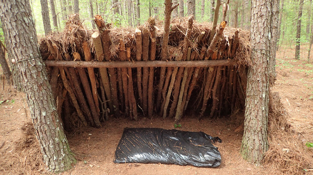 Teaching Kids Wilderness Survival: This is a lean-to shelter made from a ridgepole lashed between two trees and support poles leaning against it. Small sticks and debris are layered to create thatching for rain and wind.