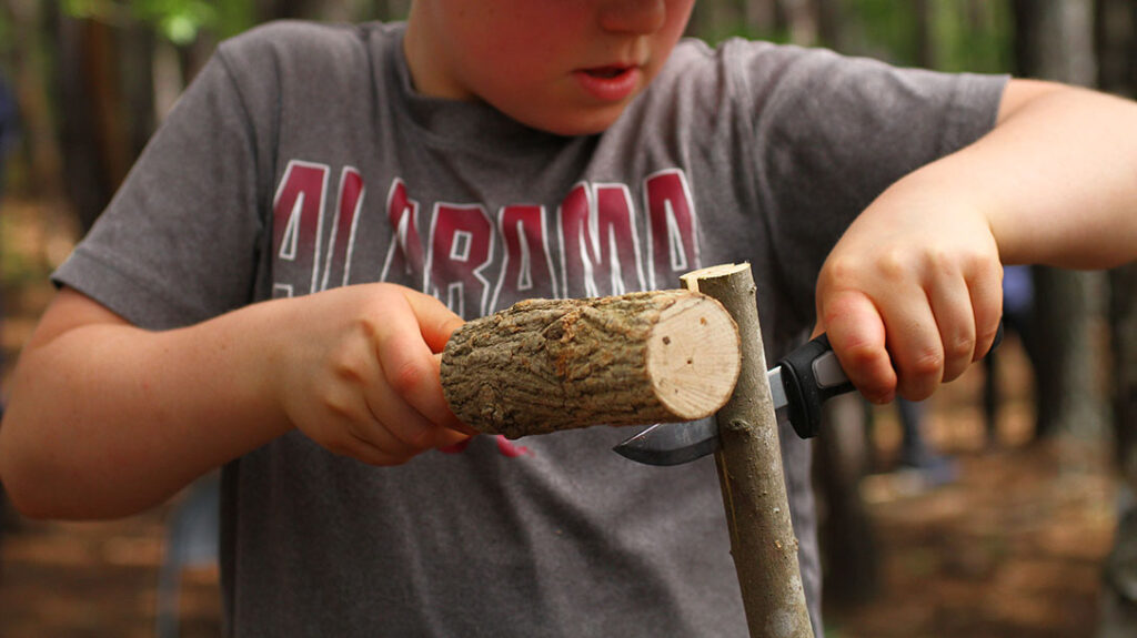 A student uses a baton and knife to split his pole, which will become a fish spear.