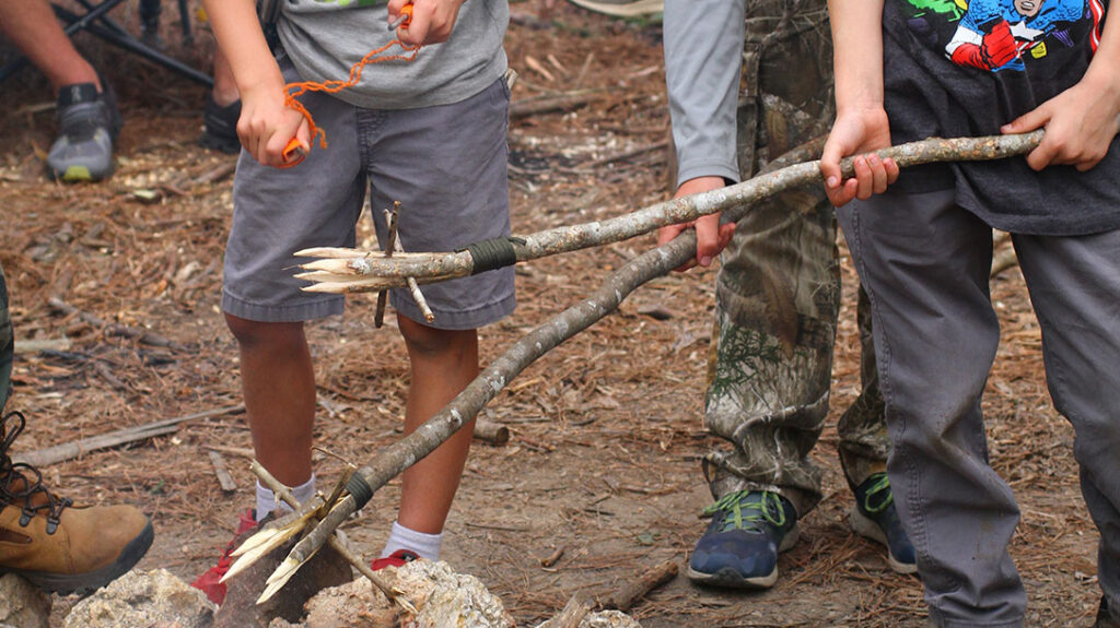 Teaching Kids Wilderness Survival: Kids are hardening their fish gigging spears near a fire. They all used a knife and cordage to complete their project.