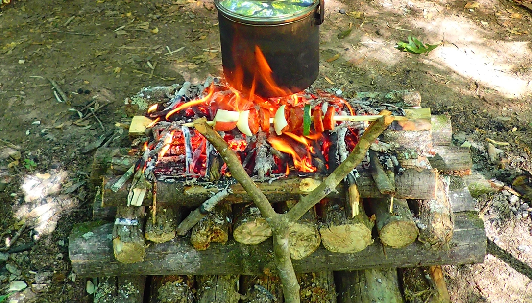 The author roasts a kabob on a rotating Y-branch over a pyramid fire while boiling a kettle, emphasizing the importance of creativity in outdoor cooking.